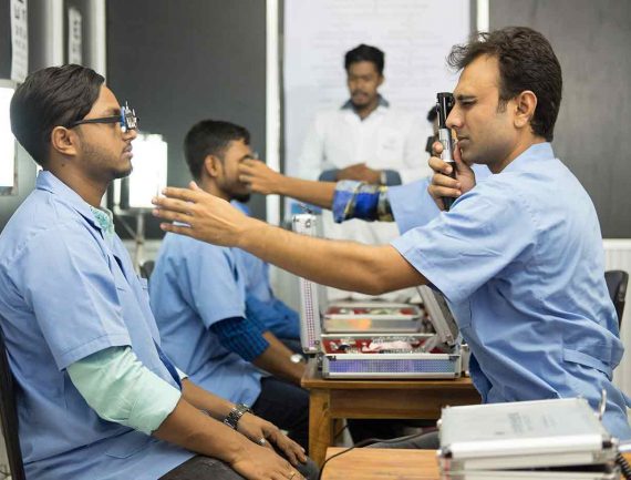 Man in blue shirt examines patient's eye with ophthalmoscope while another man in white shirt watches in background during eye exam.