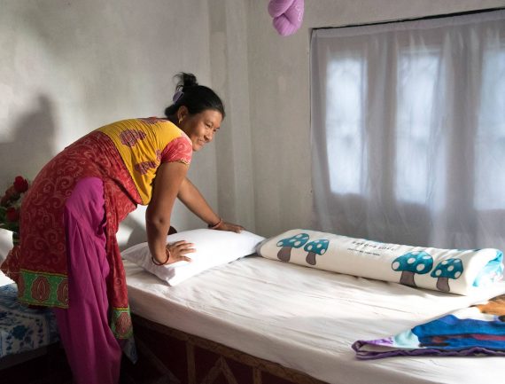 Woman in red and yellow dress making bed with white sheet, pillow, mushroom-patterned blanket, and blue and purple blanket in simple room.