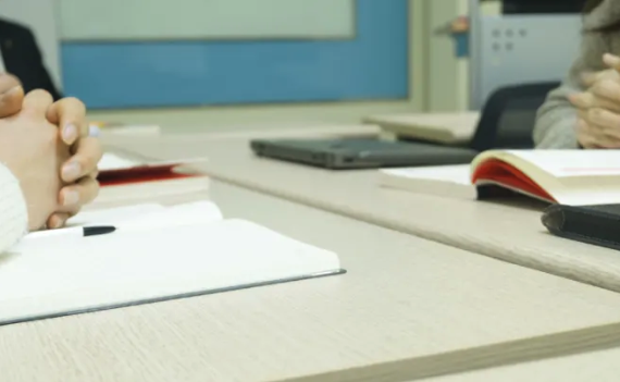 Close-up of conference table with notebook, pen, hands clasped in front. Person's face out of frame; laptop, book, folder in background.