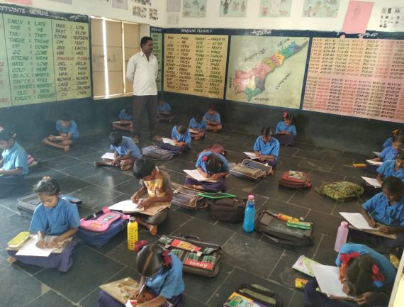 Students in blue uniforms writing as teacher instructs, with educational posters on yellow walls.