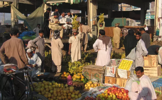 Outdoor market in South Asia with fruits and veggies on display, customers and vendors interacting, and a bicycle. Reflects street market culture in Pakistan.