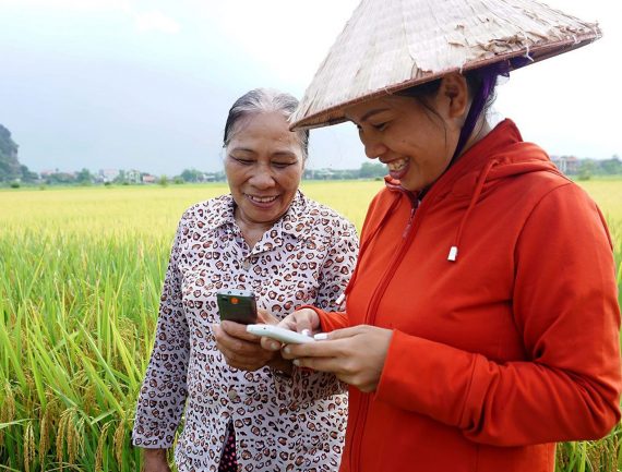Two women in a rice paddy, one in a conical hat, use a smartphone. One in a red hoodie, the other in animal print.