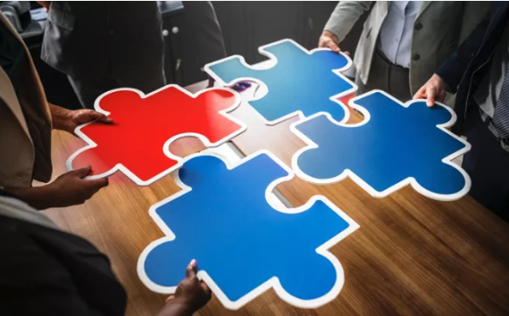 Four people collaborate around a table, each holding puzzle pieces symbolising teamwork. Red and blue pieces fit together on a wooden table surface.