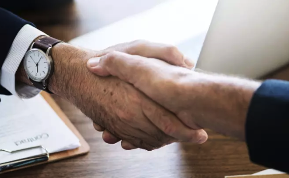 Two people shake hands over a wooden table. One person is wearing a watch.