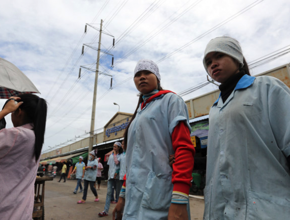 Three women in work uniforms walk in market with power line in background. One shields eyes from sun; two look ahead. Vibrant umbrella at left.