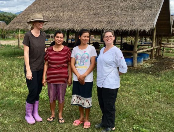Group of women in different attire standing in front of a thatched roof structure.