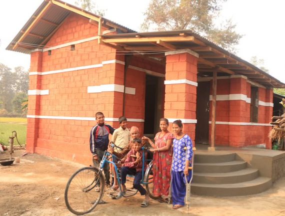 Family in front of new home, man on blue tricycle, red brick house with white trim, hand pump in background.