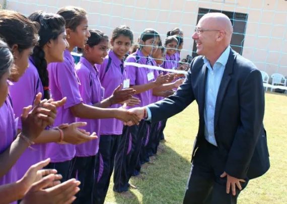 Young girls in purple shirts and black pants cheer and clap while a man in a suit shakes hands with one of them in front of a volleyball net on a grassy field.
