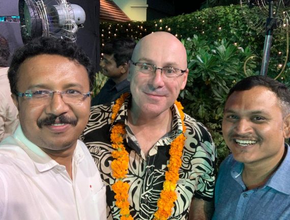 Three men posing for a photo, one wearing a lei of orange flowers and glasses, others in various shirts.