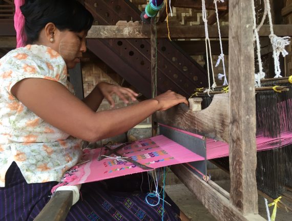 Woman weaving pink fabric on wooden loom with colorful threads, focused on her work. Hands moving quickly. .