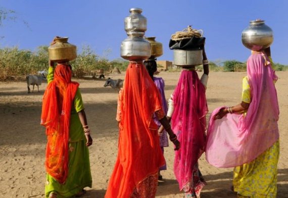 Four women in colourful saris with pots on heads walk through desert.