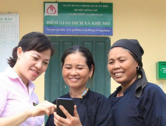 Three women interacting outside a government office, one woman pointing at her phone.