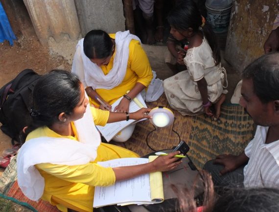 Group working in rural area, woman in yellow using solar panel to charge phone.
