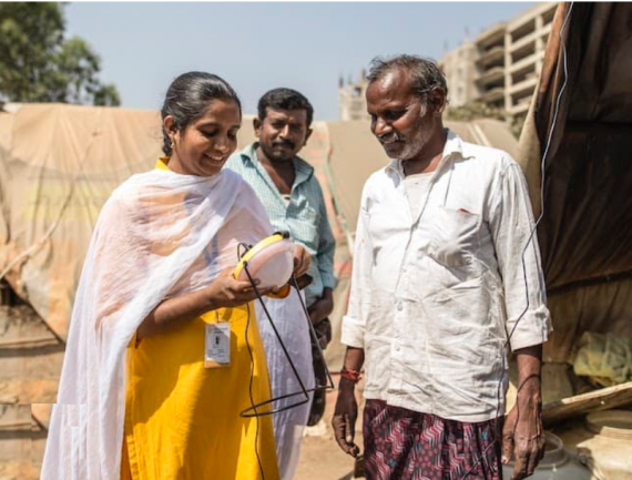 Woman in white and yellow dress with solar-powered lamp stands outdoors with two men in front of a simple makeshift home.