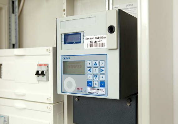 White electrical panel with black meter displaying digital readings and a label "Eigentum SWD Düren 100 069 462". Mounted with screws, second meter below main one.