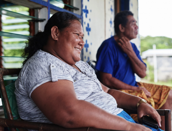 Woman smiling on porch next to man. She wears grey shirt with gold watch, he wears blue shirt. Wooden chairs and flowers on porch.