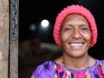 Smiling woman in pink knit hat and purple-yellow shirt stands by wooden door with metal chain.