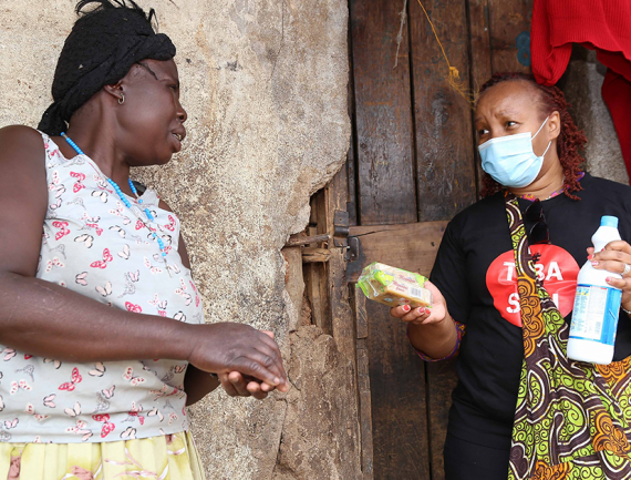 Woman in black shirt with red heart and blue mask gives soap and bleach to woman in floral dress against weathered wall.