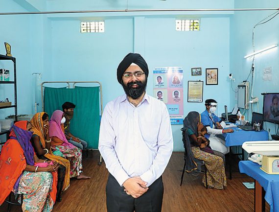 Doctor in white shirt and blue turban in clinic with patients, others in background. Blue and white color scheme.