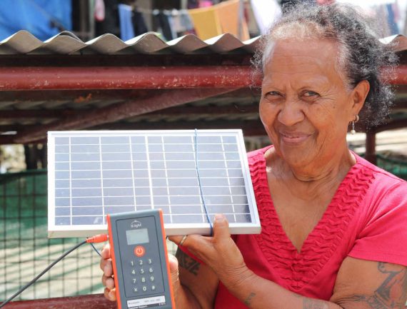 Woman with short dark hair smiles, holds up a solar panel and small device with keypad and display.