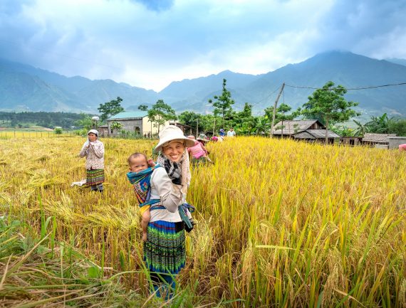Woman in traditional clothing carrying baby in sling, smiling and waving in rice field with mountains. Other women and children work in field.