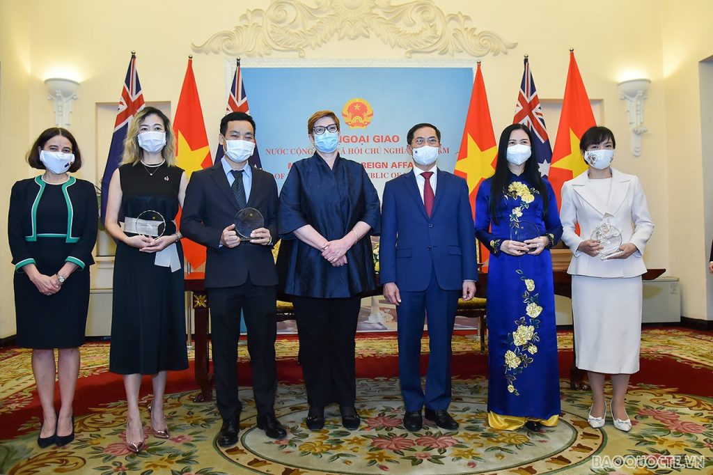 Seven people, 4 women & 3 men in masks holding plaques stand in front of flags (Australia & Vietnam). Background has "Ngo?i Giao" & "Foreign Affairs" text with tiled floor.