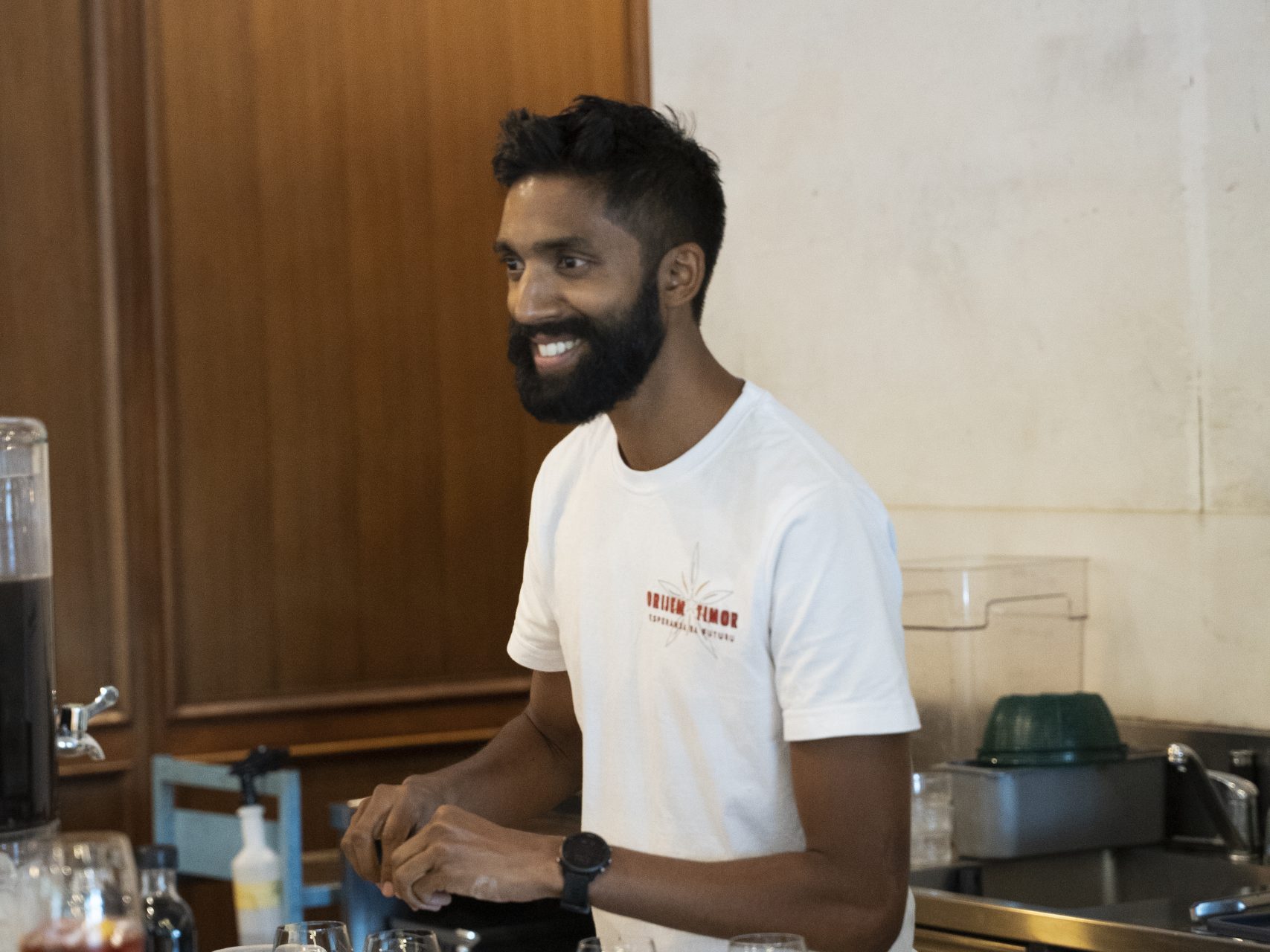 Man in white tee with "Orlaism Timor" logo making drinks at a bar, red wine glass in front.