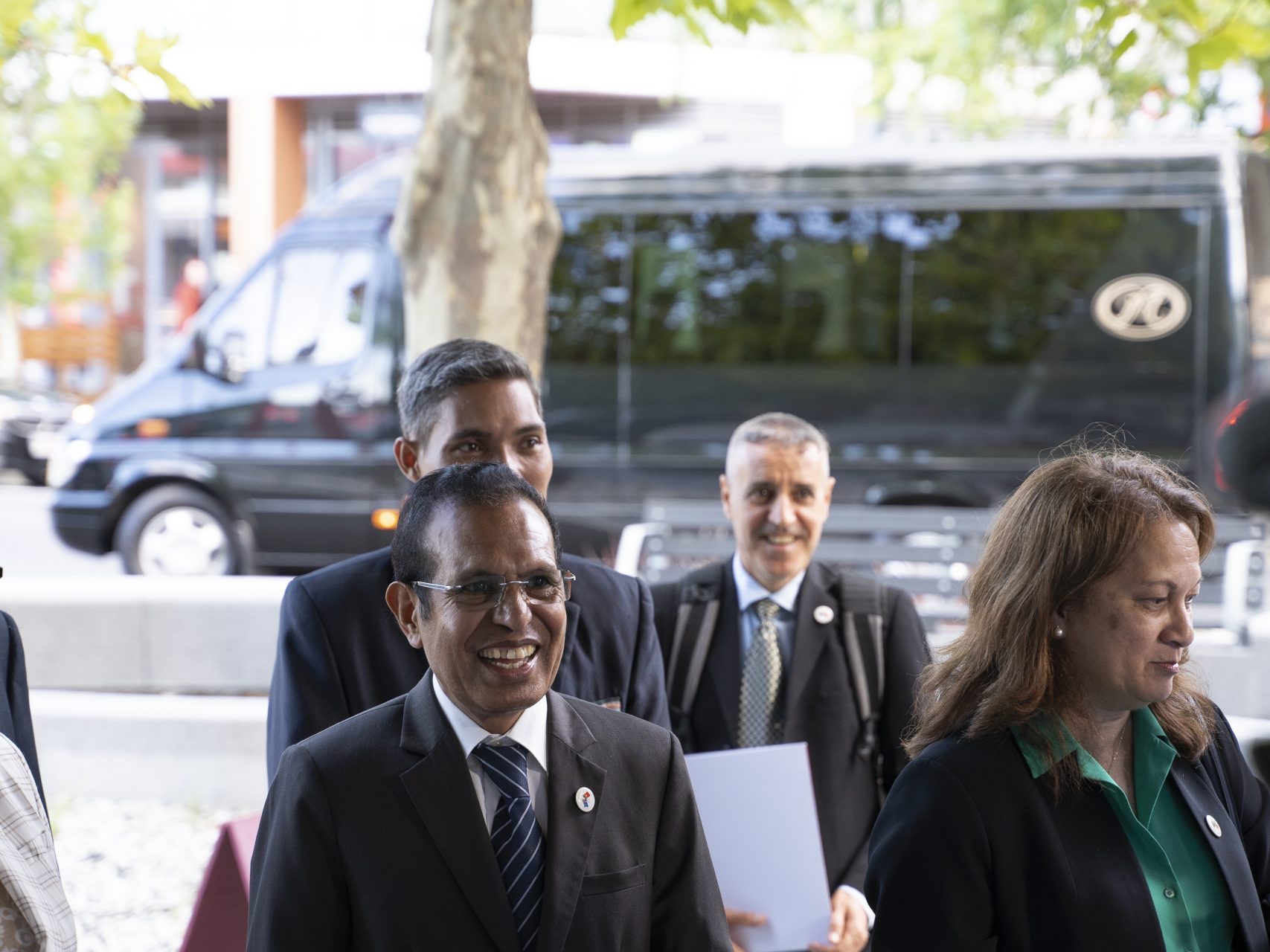 Four people, three men, and one woman in front of a black van. The central man is smiling in a black suit, blue and white tie, and glasses, surrounded by colleagues and nature.