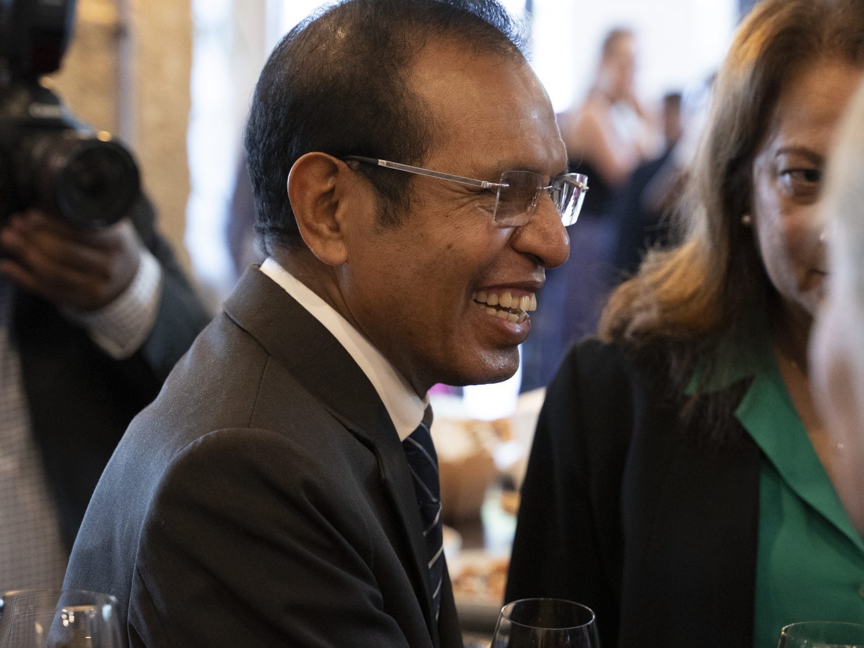 Man in suit and glasses smiling with red wine in a crowded room, talking to a woman in green shirt. Background shows camera and part of another person.