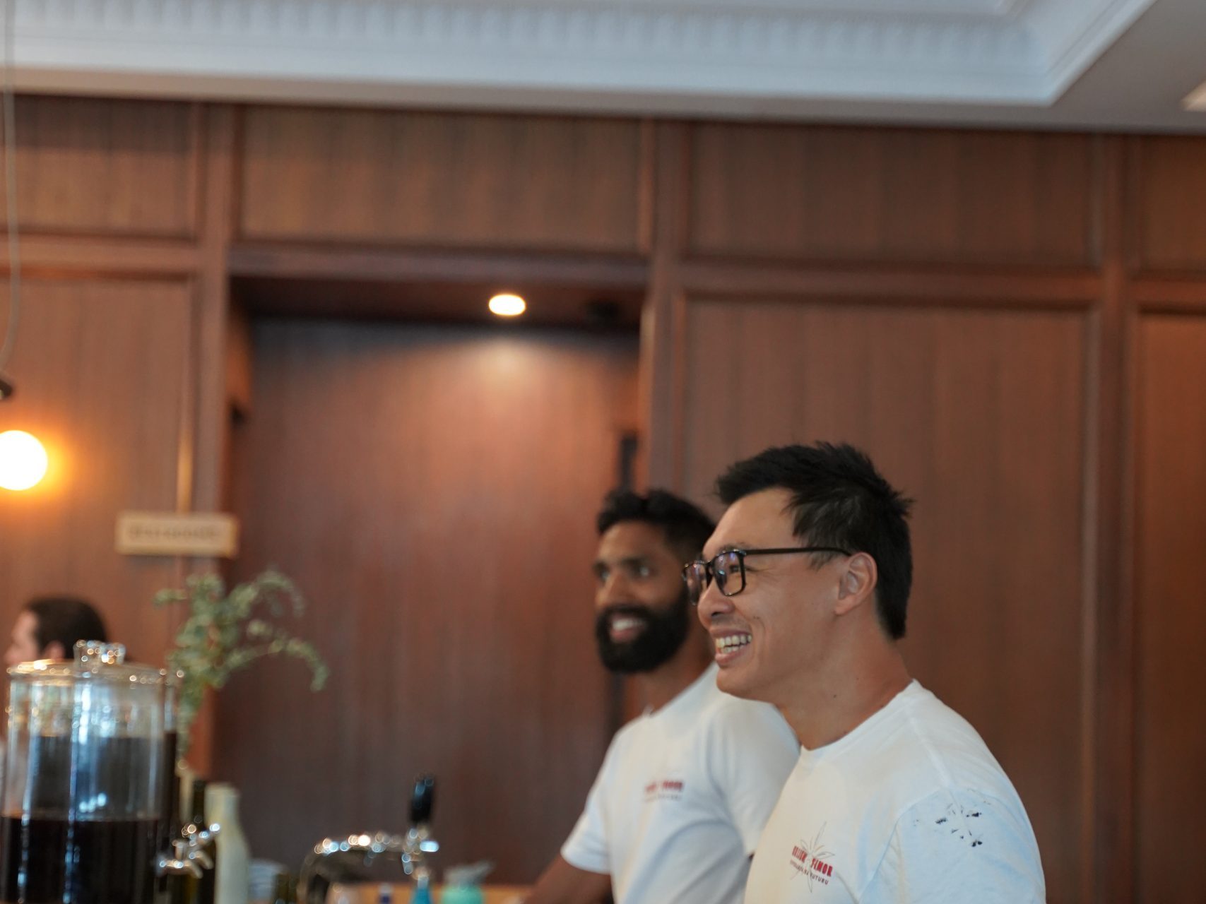 Two men at a wooden bar, wearing white t-shirts, glasses of wine and a red pitcher in front.