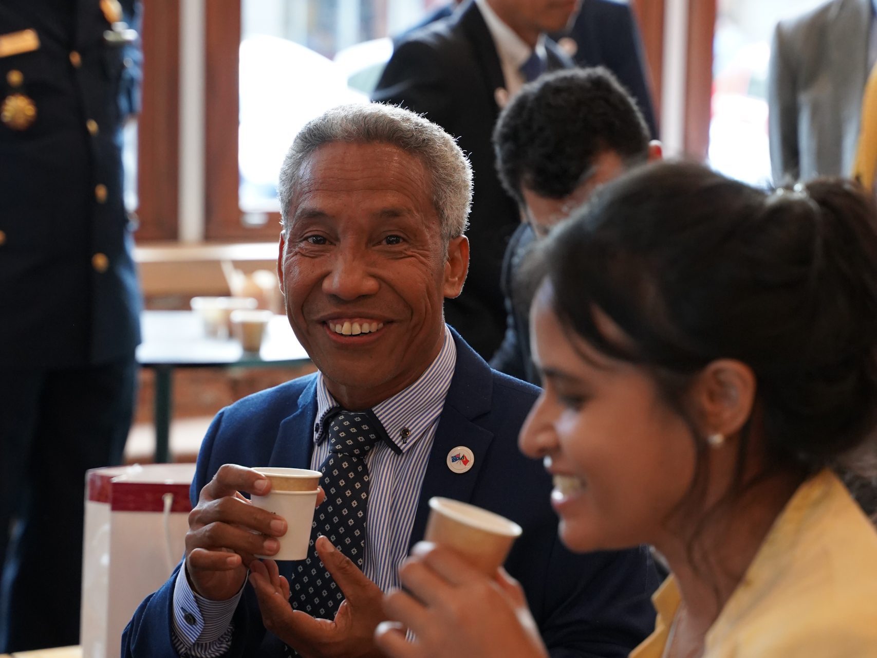 A man in a suit smiles holding a coffee cup while a woman in a yellow jacket holds a coffee cup beside him. Another man in a suit is blurred in the background. The man in the suit has a flag pin on his jacket. The woman in the yellow jacket is smiling.