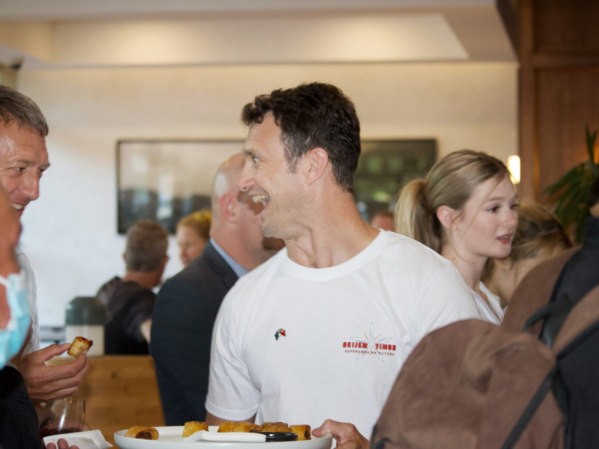 Man in white shirt with "Orijen Timor Esperanca ba Futuru" holding pastries indoors.