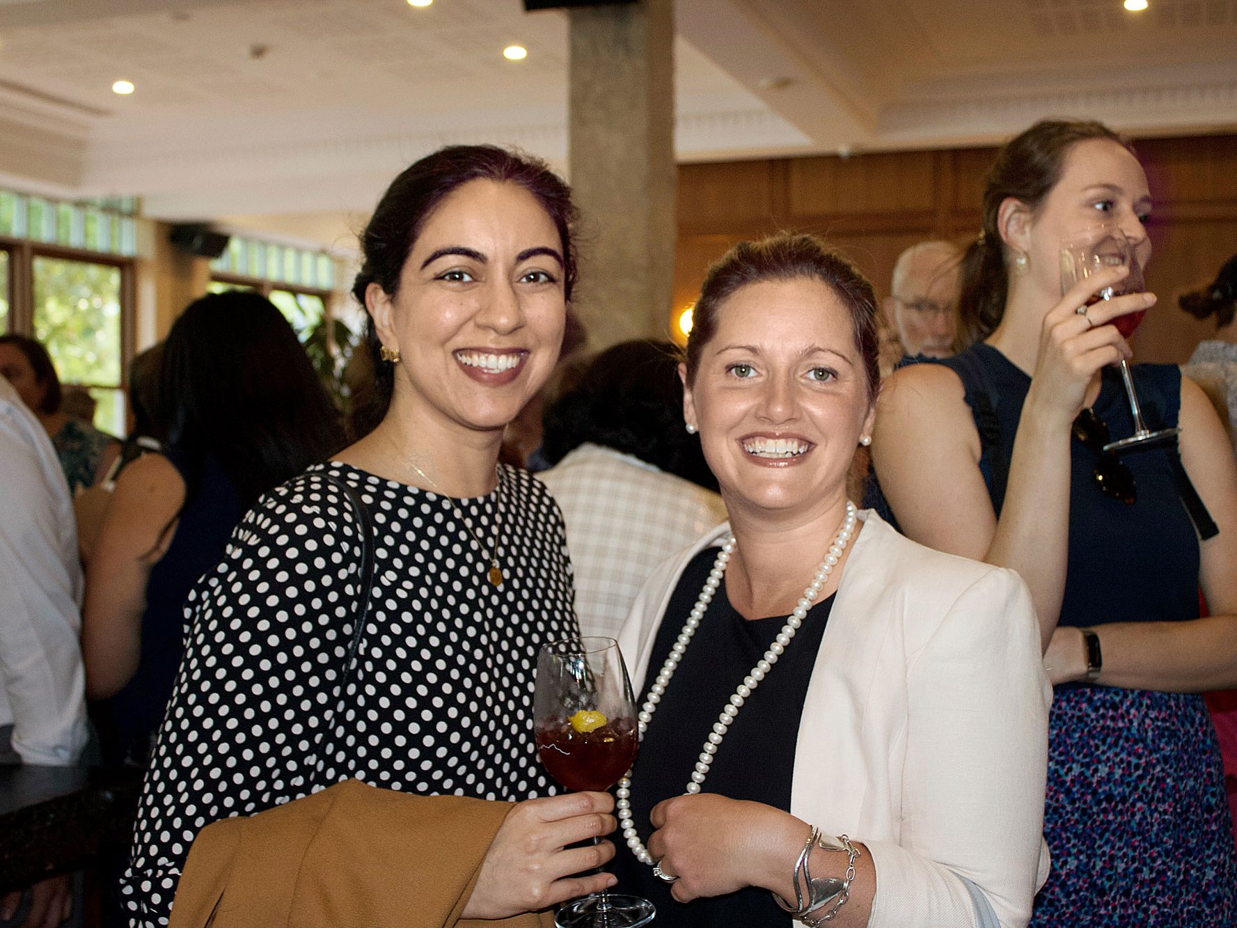 Three women smiling and holding wine glasses at a social event. One woman in a white blazer and pearl necklace, another in a polka dot shirt and brown jacket, and the third in a blue top and patterned skirt, surrounded by others.