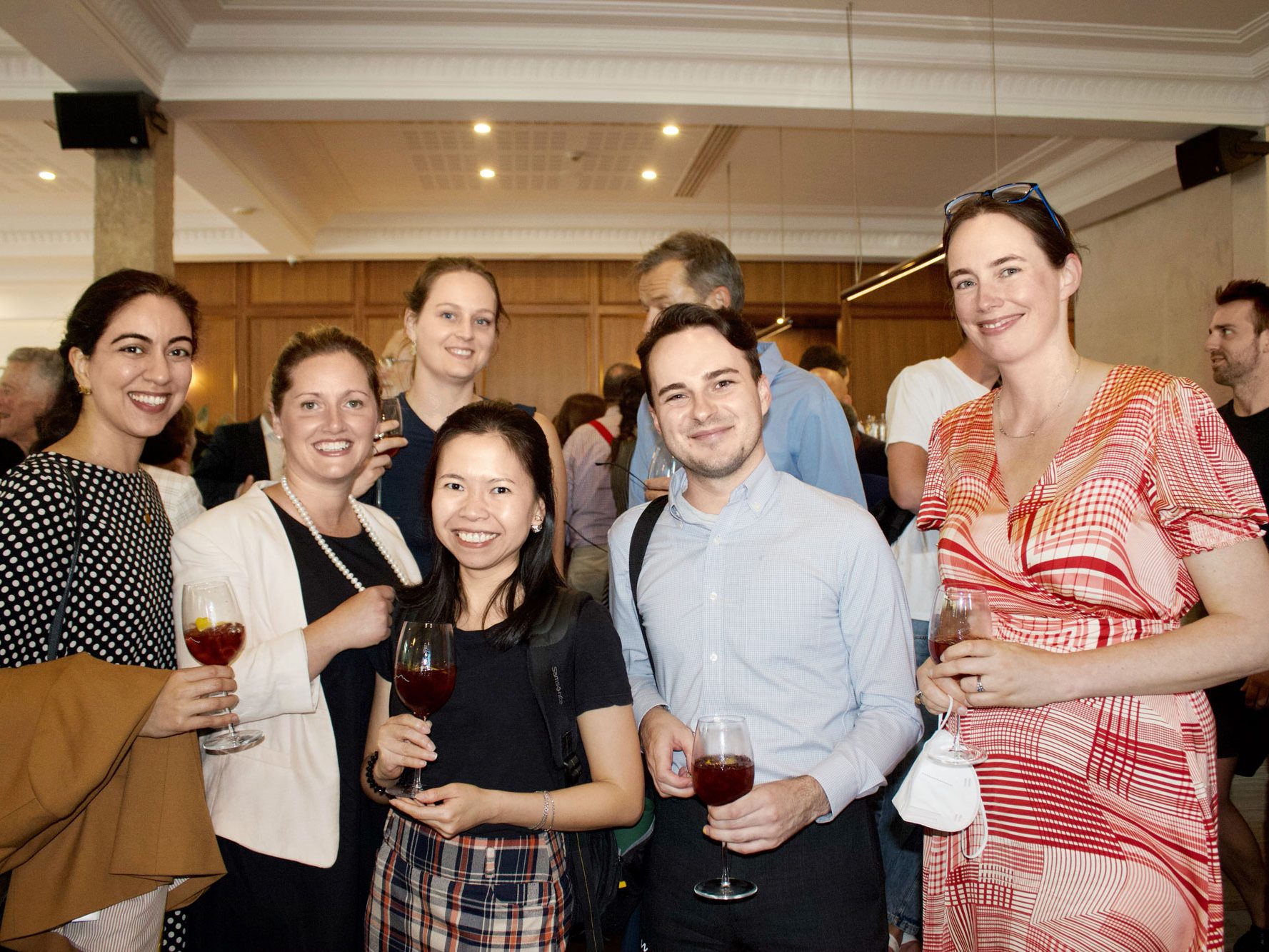 Friends at a bar, smiling with glasses of wine. Woman in red and white dress holds a white face mask.