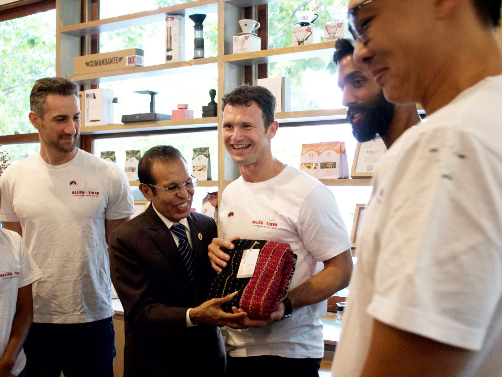 A group celebrating in a coffee shop, one man hands a wrapped gift to another, all smiling.